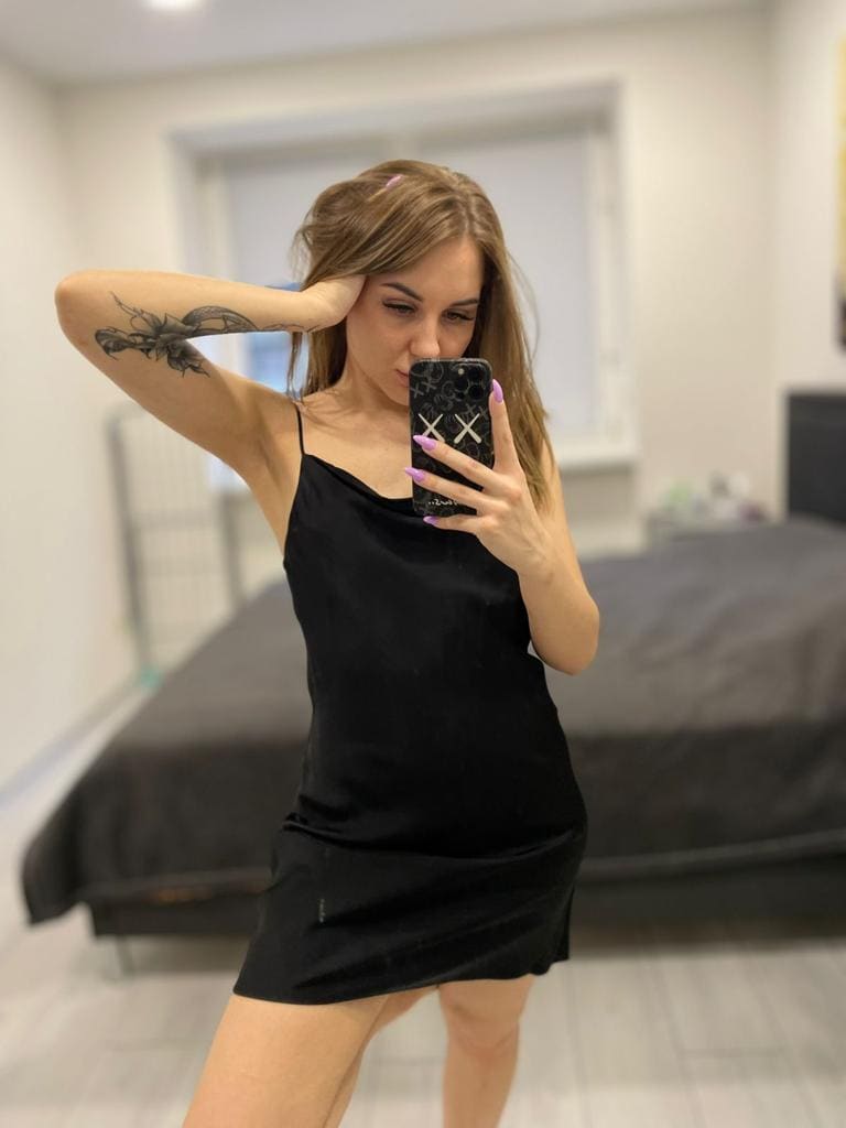 A brunette with a floral arm tattoo takes a mirror selfie in a bedroom, wearing a short black slip dress with thin straps.