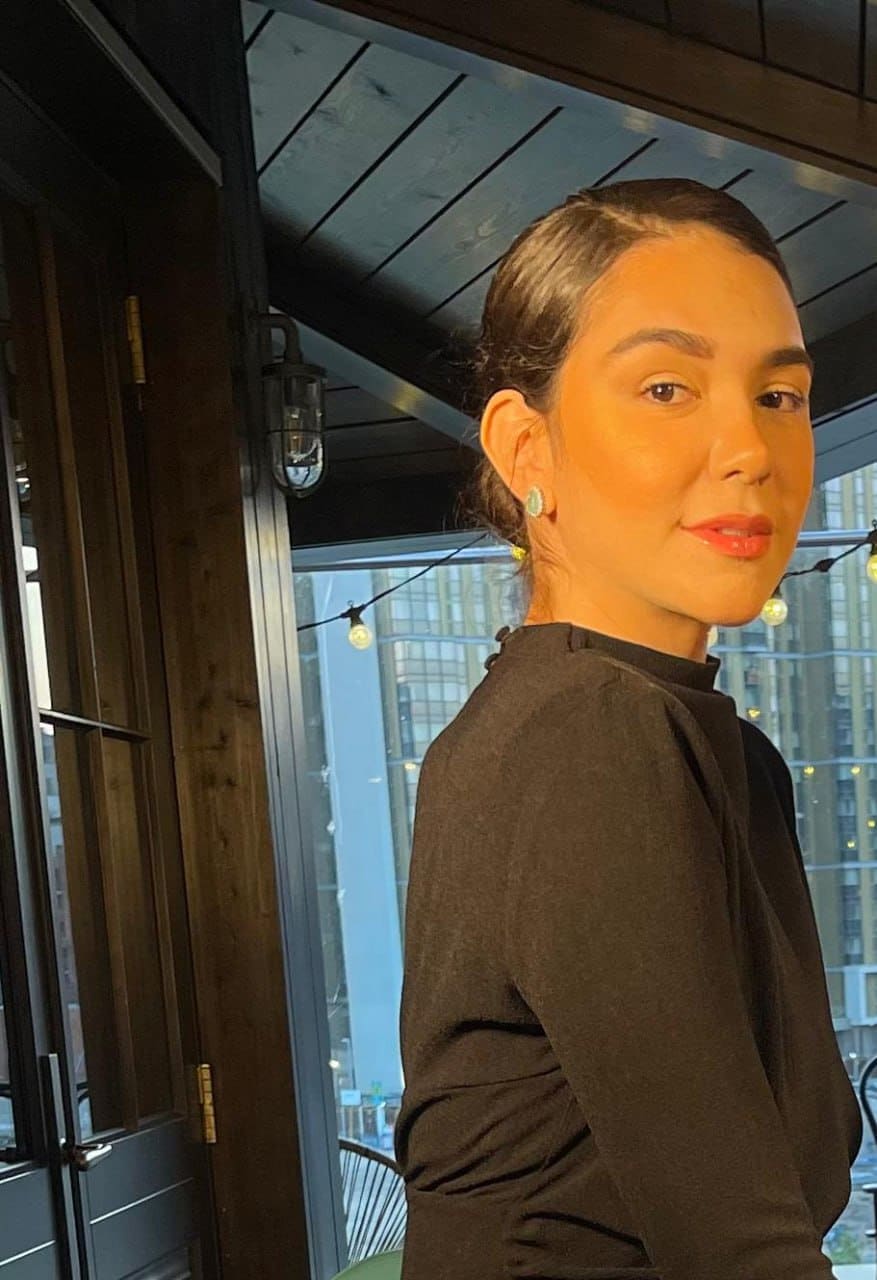A woman in a black high-neck top stands in profile in a room with dark wood paneling and string lights outside.