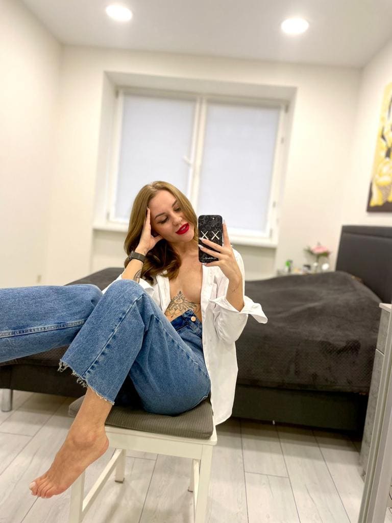 A barefoot woman sits on a white stool in an open white shirt and denim overalls, holding a phone and resting a hand on her head.