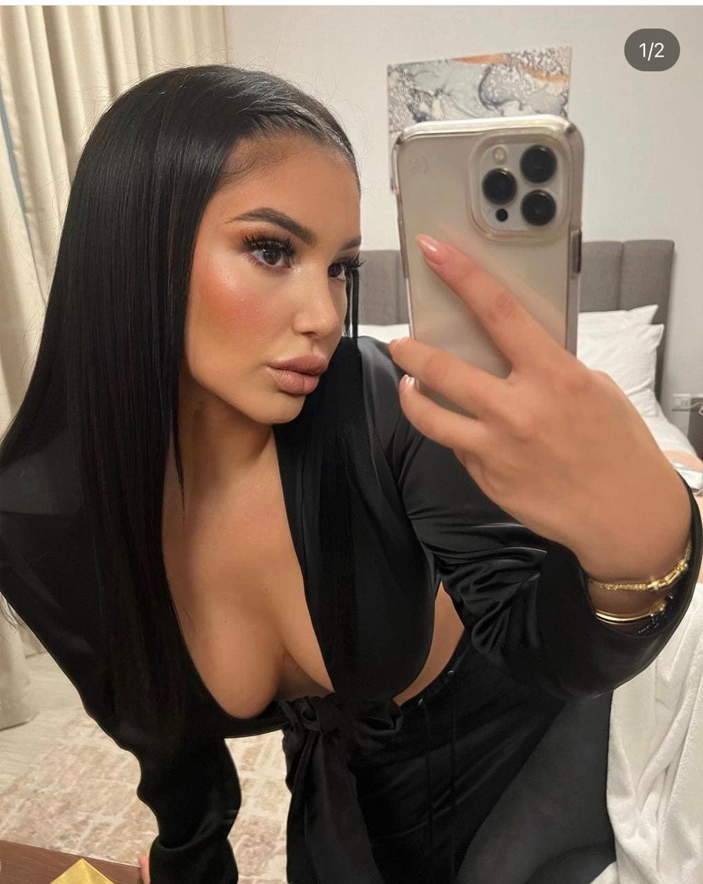 A woman takes a mirror selfie in a bedroom, wearing a black long-sleeved top with a deep neckline and side cutout.