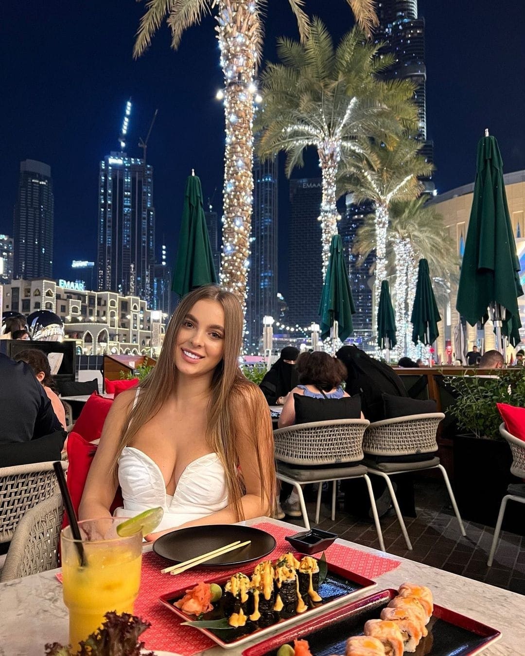 Janette sits at an outdoor restaurant table at night in a white strapless top, with sushi plates and a city skyline behind her.