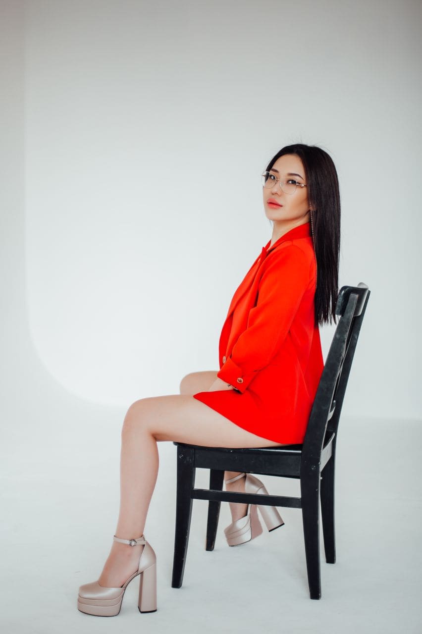 Lisa sits on a black chair in a white studio, wearing a red blazer dress and beige platform heels, looking over her shoulder.