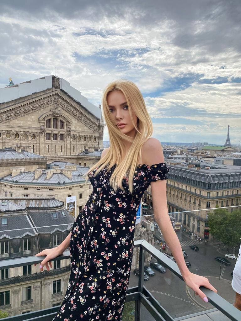 Victoria stands on a balcony railing in a black floral off-shoulder dress, with the Eiffel Tower and city skyline behind her.
