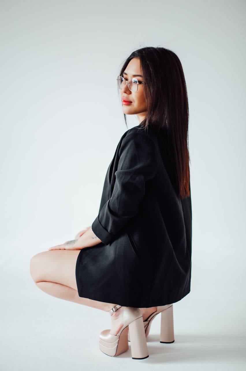 A woman crouches in profile against a white background, wearing an oversized black blazer as a dress and beige platform sandals.