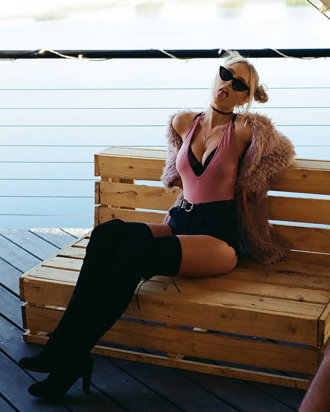 Woman with hair in a bun sits on a wooden pallet bench by water, wearing a pink top, black shorts, thigh-high boots, and a fur coat, sticking her tongue out.