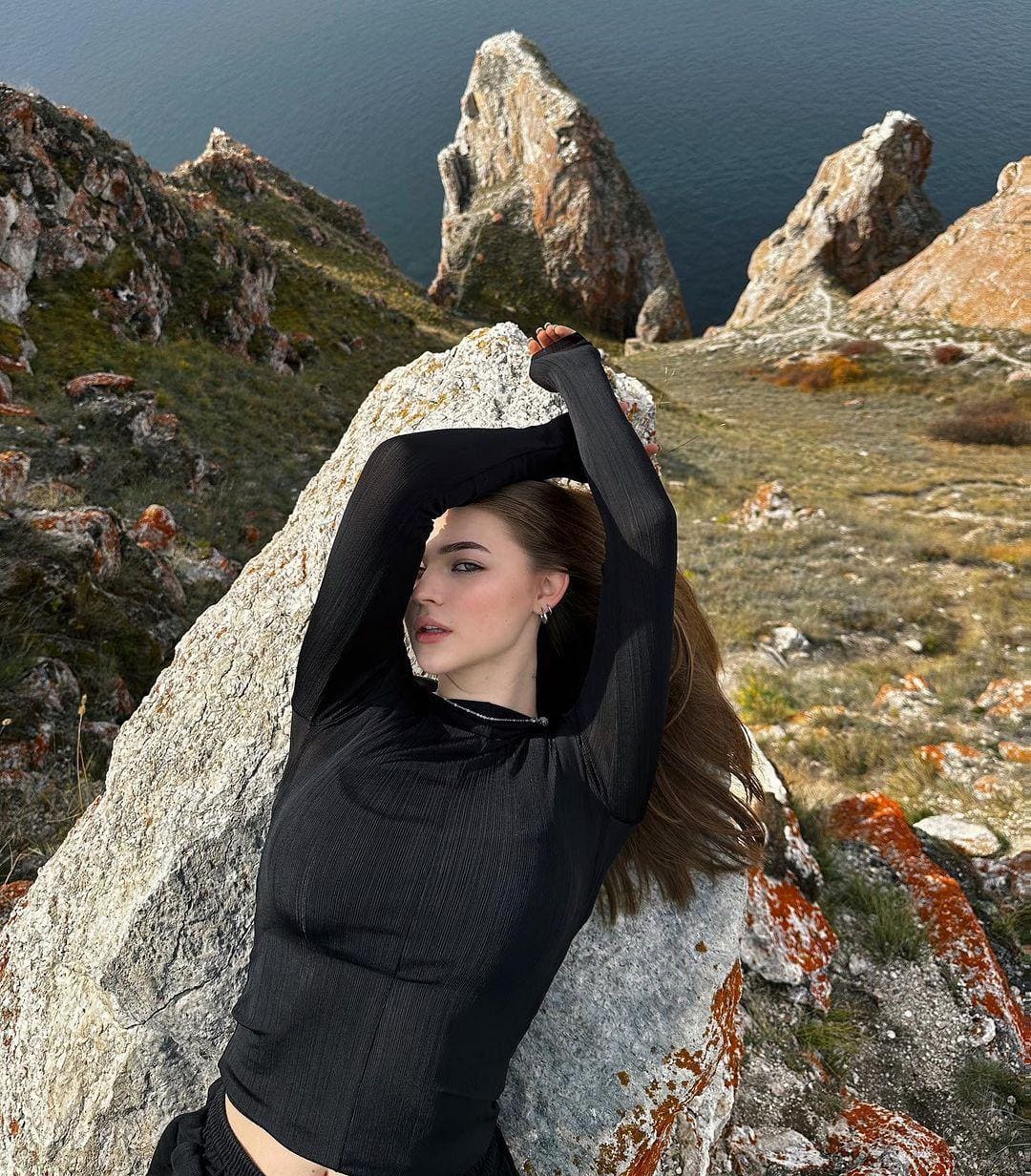 A woman with long brown hair stands on a rocky coastal cliff in a black long-sleeved top, arms raised above her head near sea stacks.