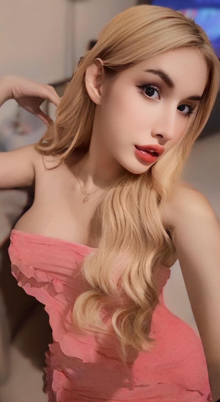 A woman with wavy blonde hair wears a pink strapless ruffled top, posing with a hand near her head against a blurred indoor background.