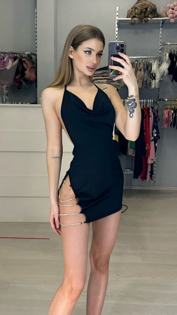 Malika takes a mirror selfie in a black halter mini dress with chain details, standing in a room with clothing racks behind her.