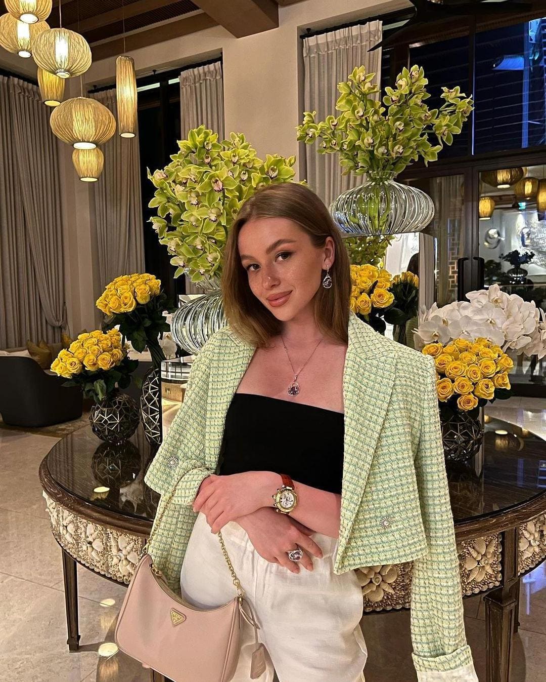 Emily stands in a flower-filled lobby wearing a green tweed jacket, black top, white trousers, and holding a pink handbag.