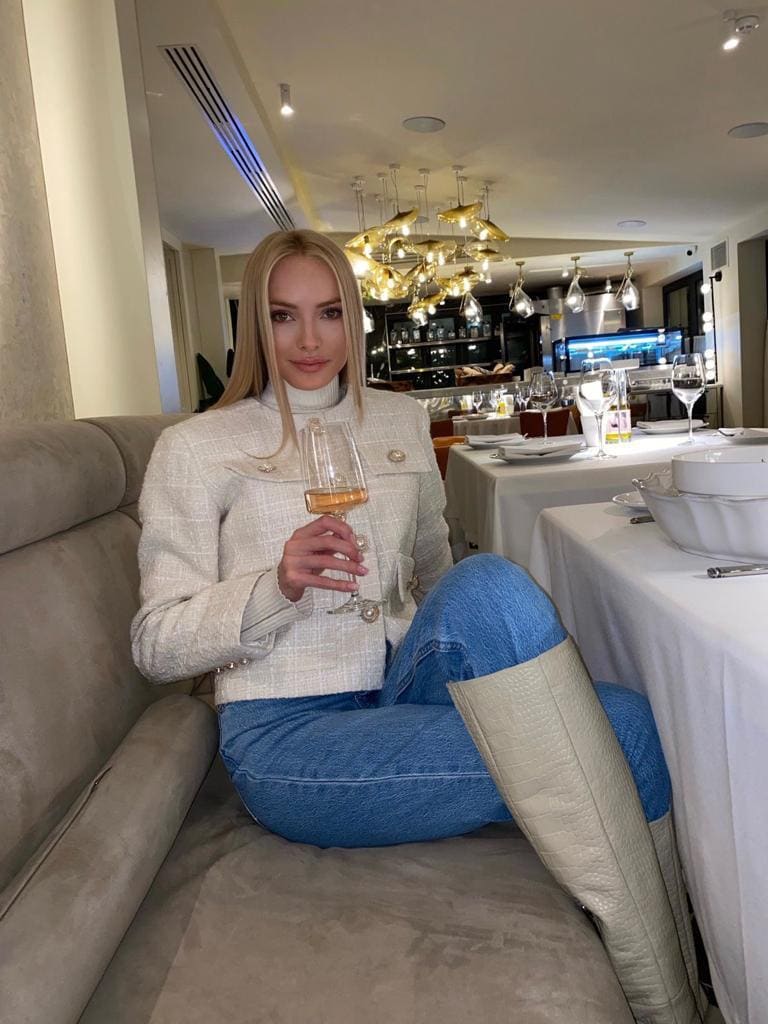 A blonde woman sits on a beige sofa in a white jacket and jeans, holding a glass of rosé wine in a restaurant setting.