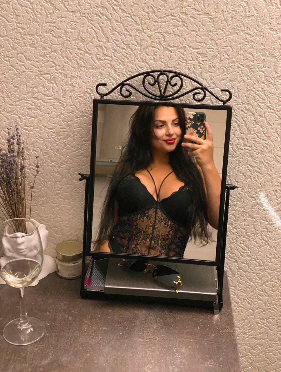 A woman in a black lace bodysuit takes a mirror selfie in front of a decorative black-framed mirror, with a wine glass and dried flowers nearby.