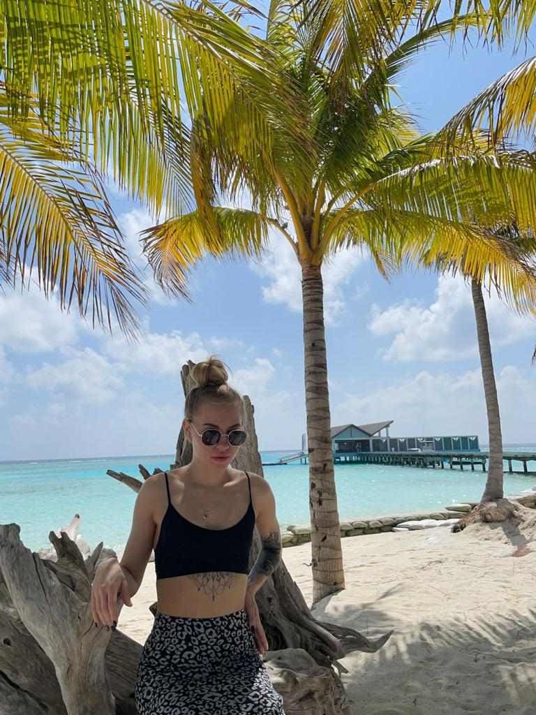 A blonde woman in a black crop top and patterned skirt stands on a beach with palm trees and turquoise water behind her.