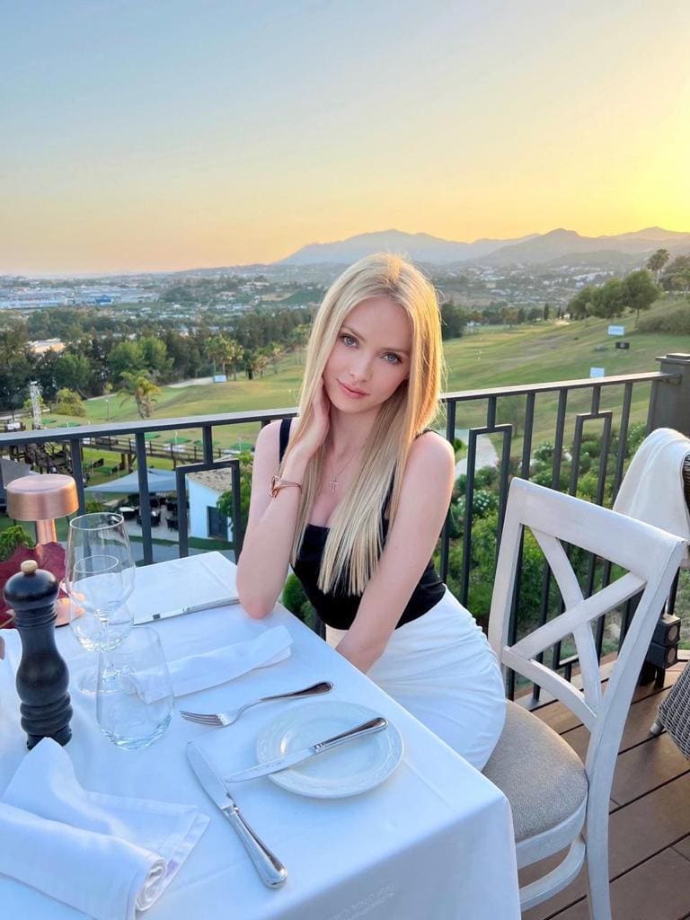 A blonde woman sits at an outdoor dining table in a black top and white skirt, resting her hand near her chin against a sunset.