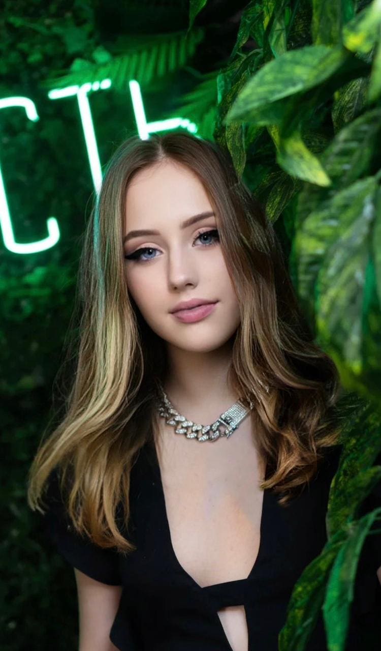 Standing before green foliage and a neon sign, she wears a black short-sleeved V-neck top and a chunky silver necklace, with wavy brown hair.