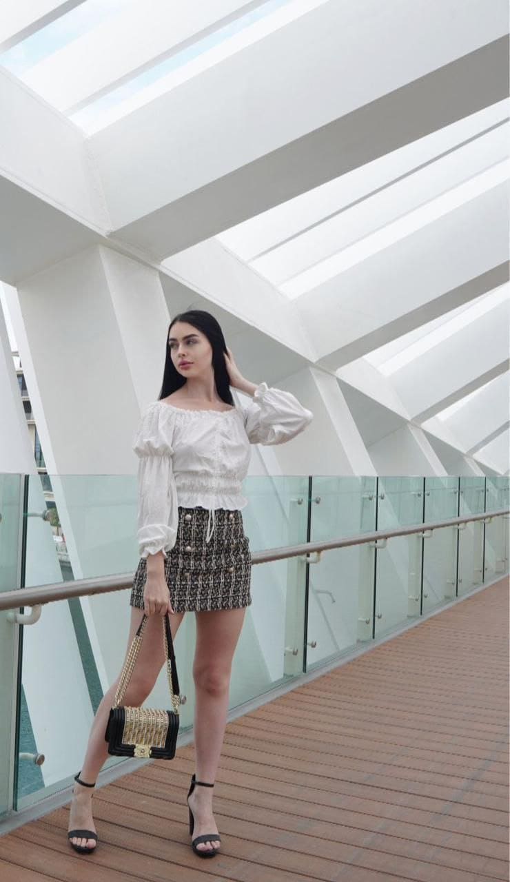 A woman in a white off-shoulder blouse and patterned skirt stands on a wooden walkway, holding a small woven handbag.