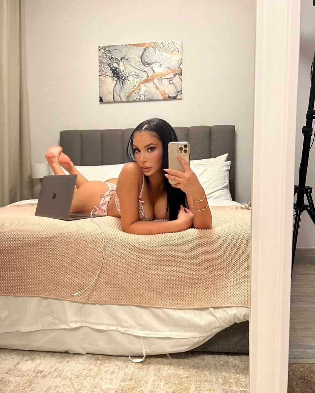 A woman in light lace lingerie lies on her stomach on a bed, holding a smartphone with an open laptop nearby.