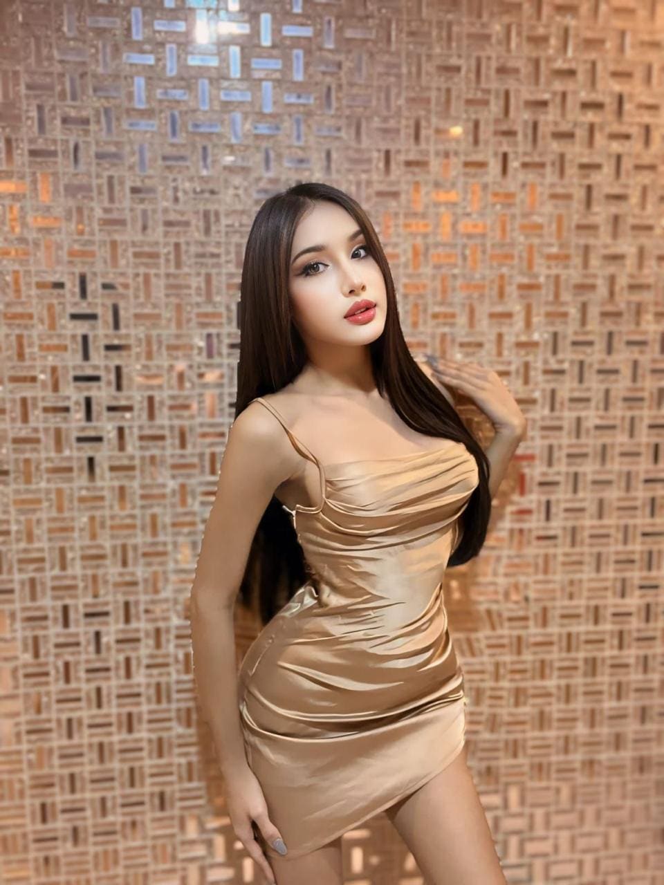 A woman in a gold satin mini dress with thin straps poses against a mosaic wall, one hand near her shoulder and the other on her hip.