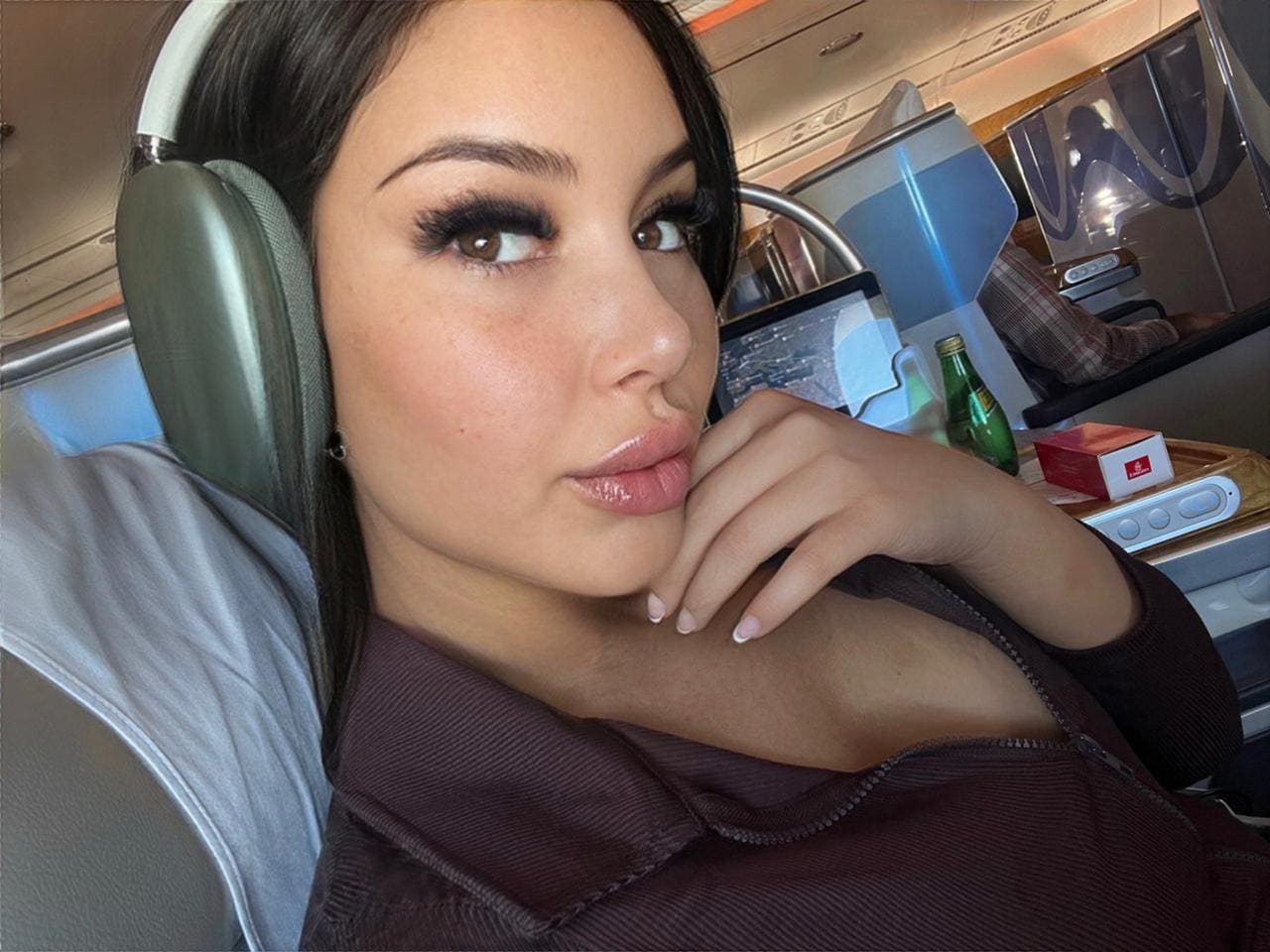 A woman sits in an airplane seat wearing large headphones and a dark zip-up top, resting her chin on her hand.