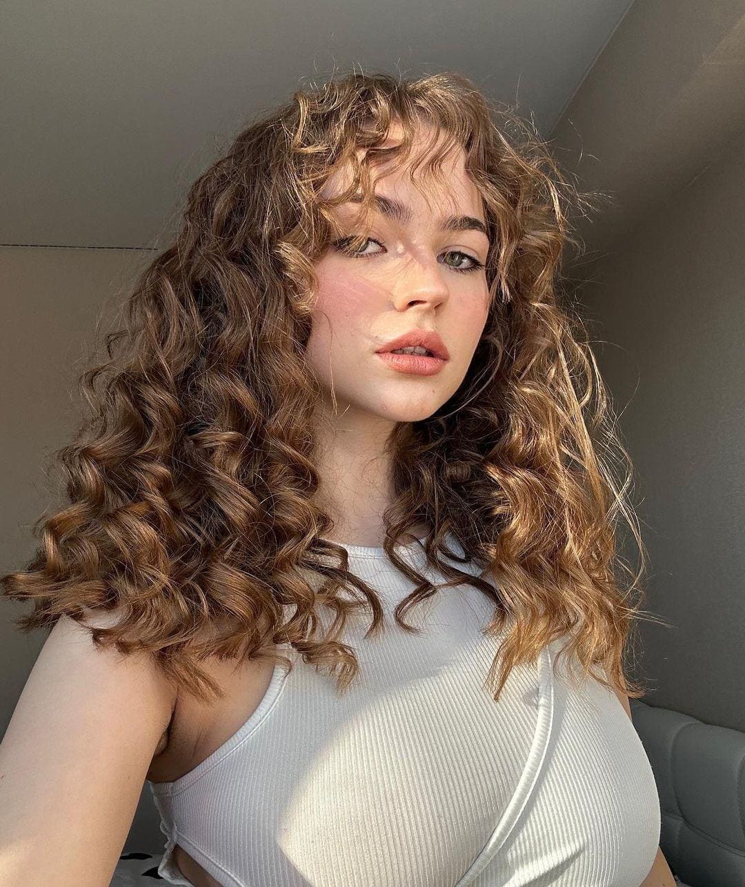 Romy with long curly brown hair wears a white ribbed tank top, posing indoors against a plain wall with sunlight on her chest.