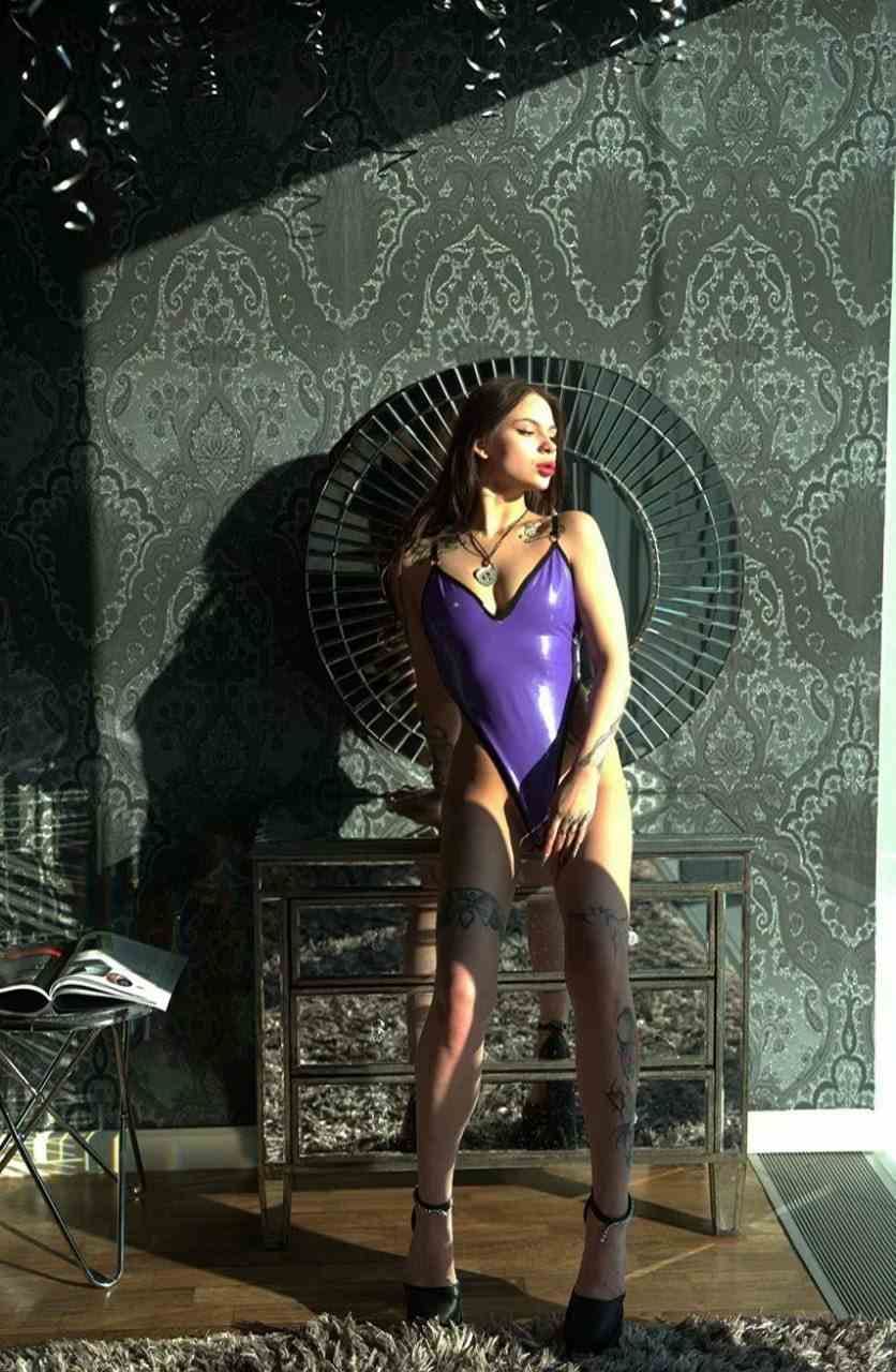 A woman stands in a purple bodysuit and black heels, facing forward near a round mirror and cabinet, against patterned wallpaper.