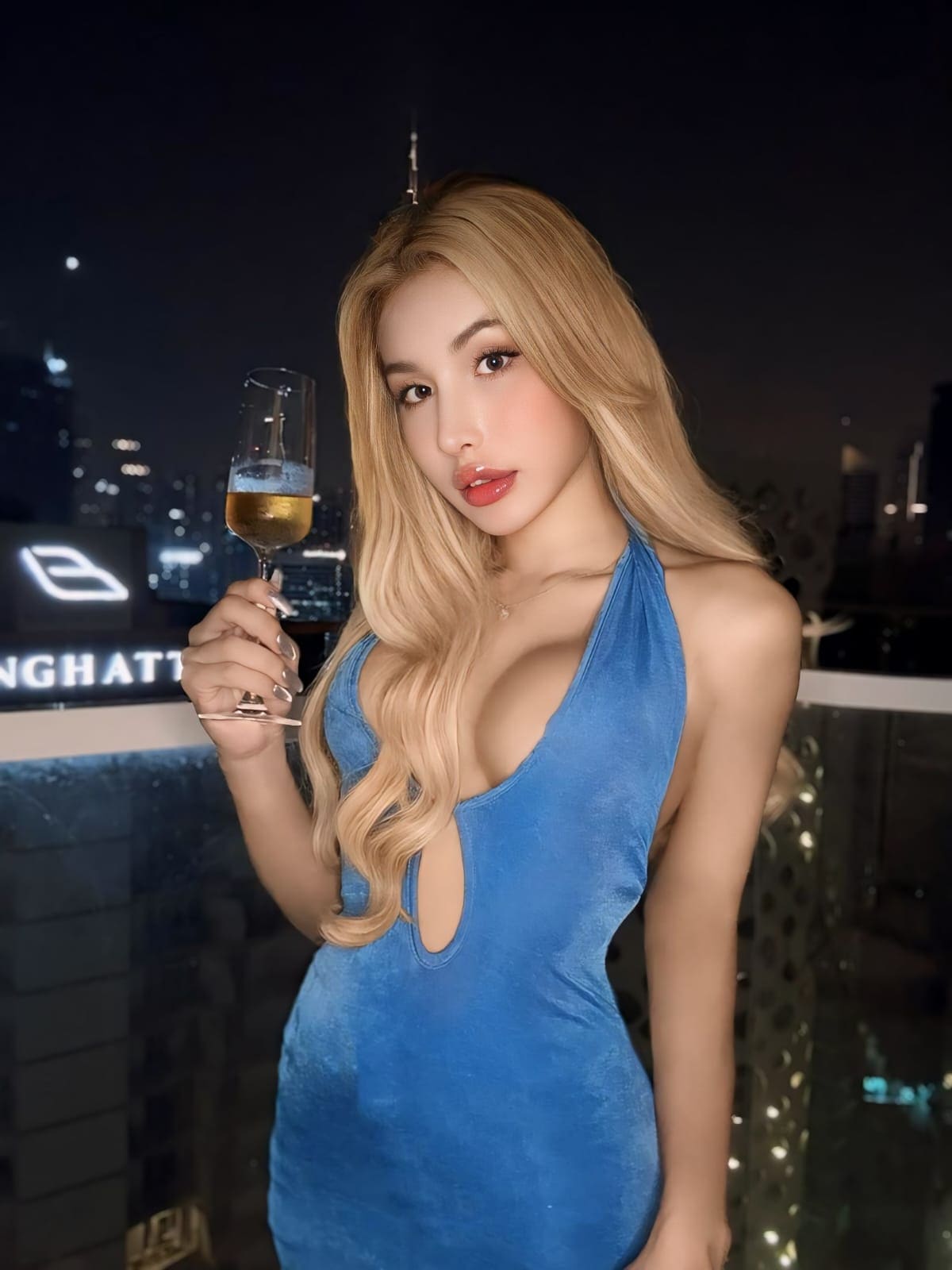 A blonde woman holds a champagne glass on a rooftop at night, wearing a blue velvet halter dress with a keyhole cutout and city lights behind.