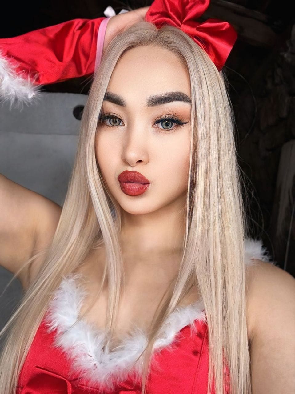 A woman with a red bow headband poses with one arm raised, wearing a red sleeveless top trimmed with white faux fur in a dim indoor setting.