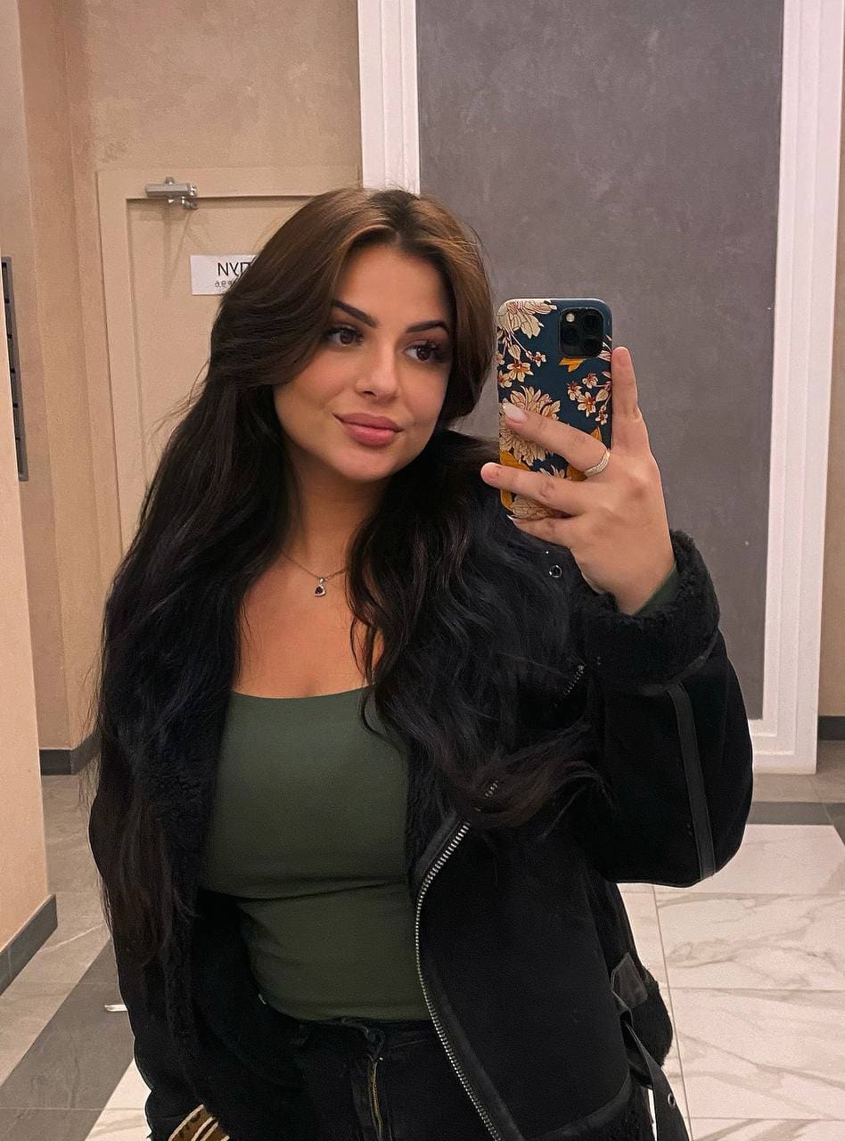 Karina takes a mirror selfie in a hallway, wearing an olive tank top under an open black fur-lined jacket, holding a floral phone case.