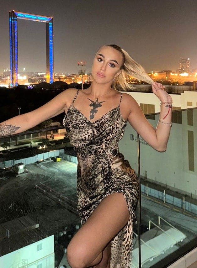 A blonde in a leopard-print dress stands on a rooftop at night, holding her hair near a railing with an illuminated arch behind.