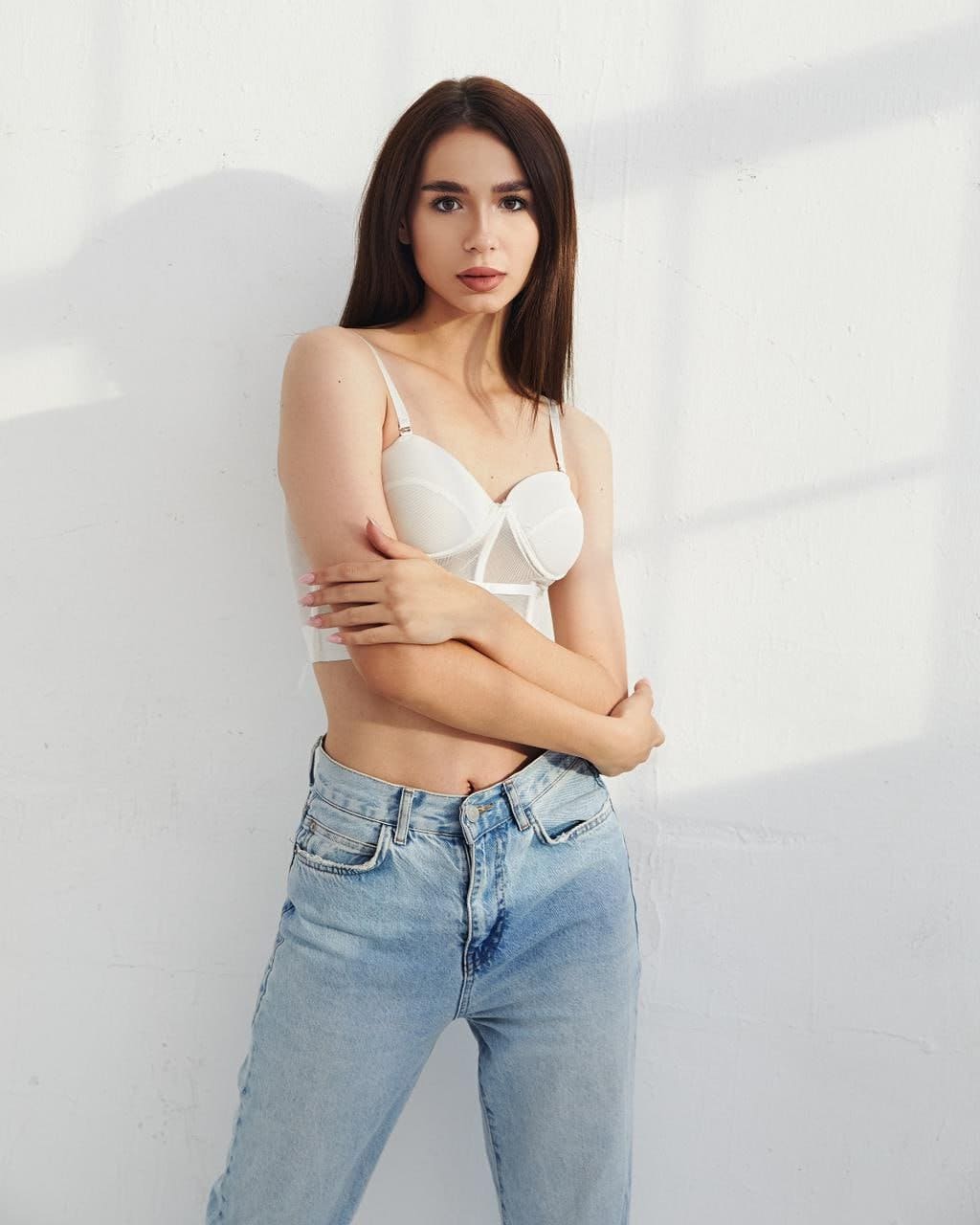 Milly stands against a white wall in a white bra and light blue jeans, arms crossed over her chest.