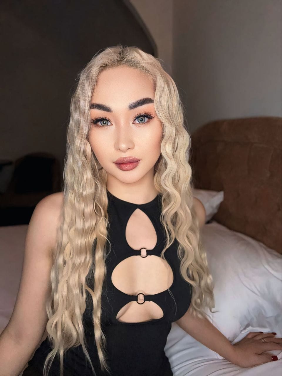 Harley sits on a bed with white linens, wearing a black sleeveless top with chest cutouts, facing the camera with her left hand on the mattress.