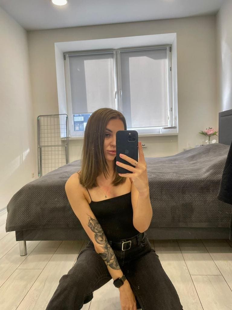 A brunette in a black spaghetti-strap top and dark jeans sits on a wood floor taking a mirror selfie with a black phone.