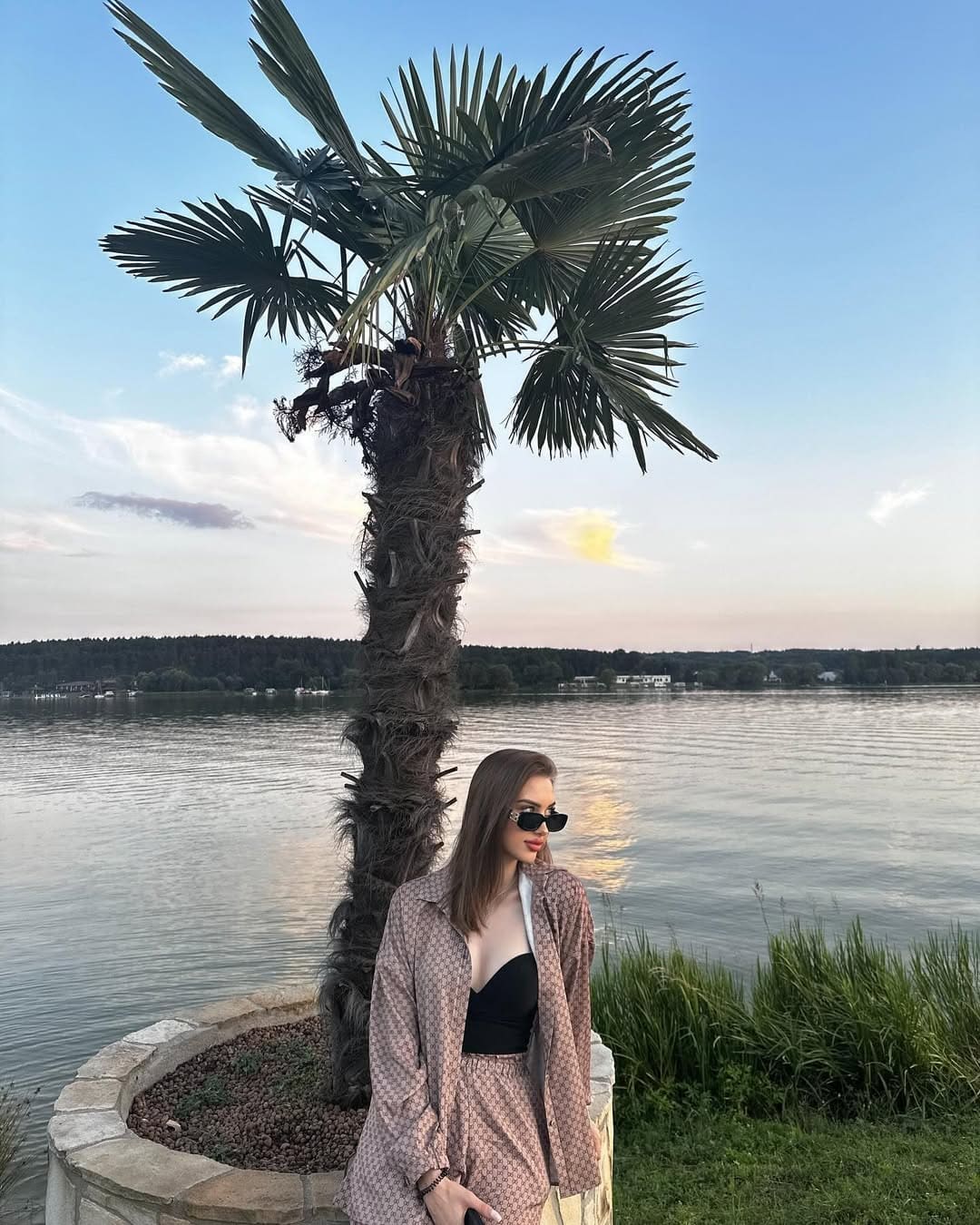 A woman in sunglasses and an open patterned shirt over a black bandeau stands by a lake near a potted palm tree.