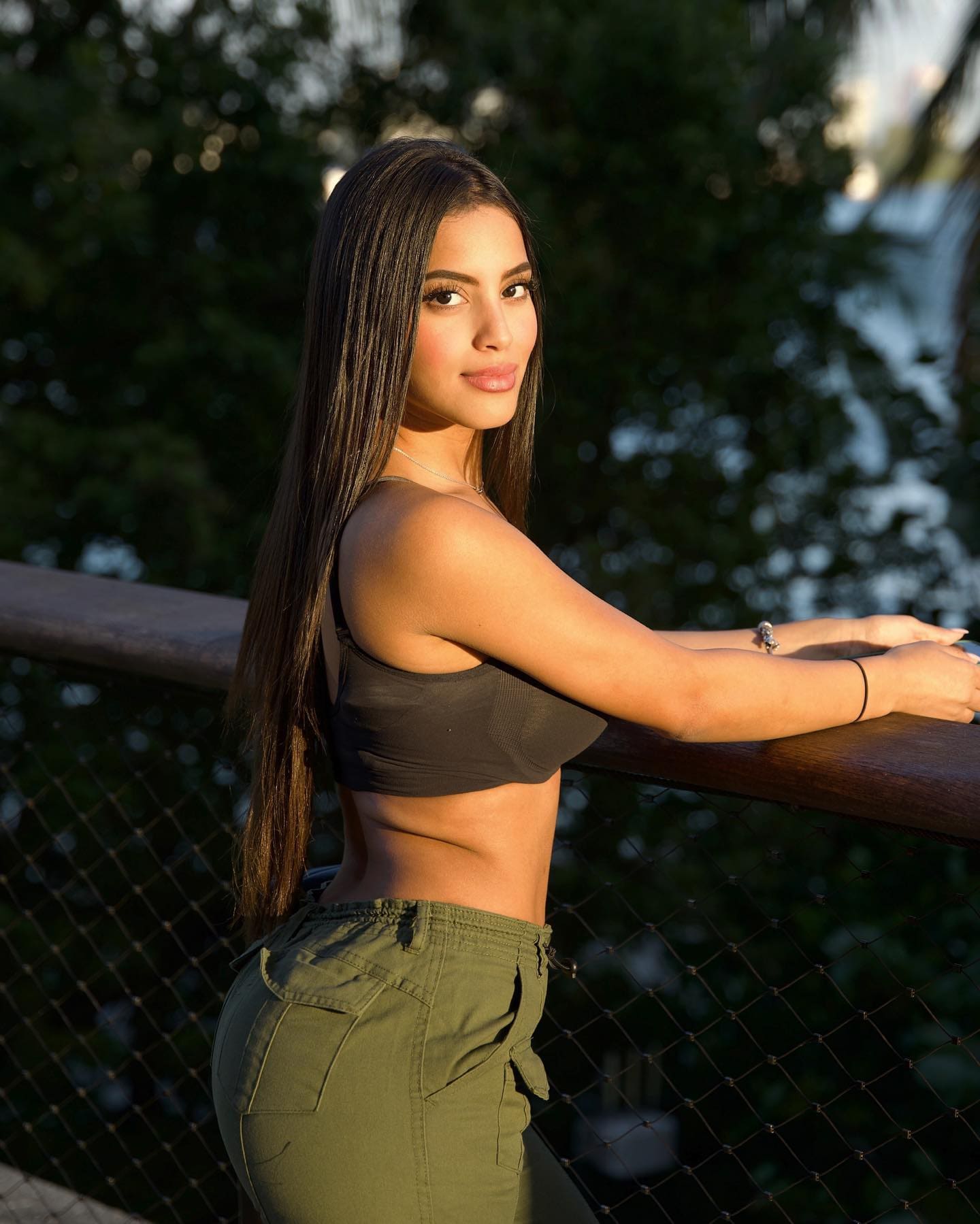 Haley stands at a wooden railing in a dark cropped tank and olive cargo pants, arms resting on the rail against blurred trees.
