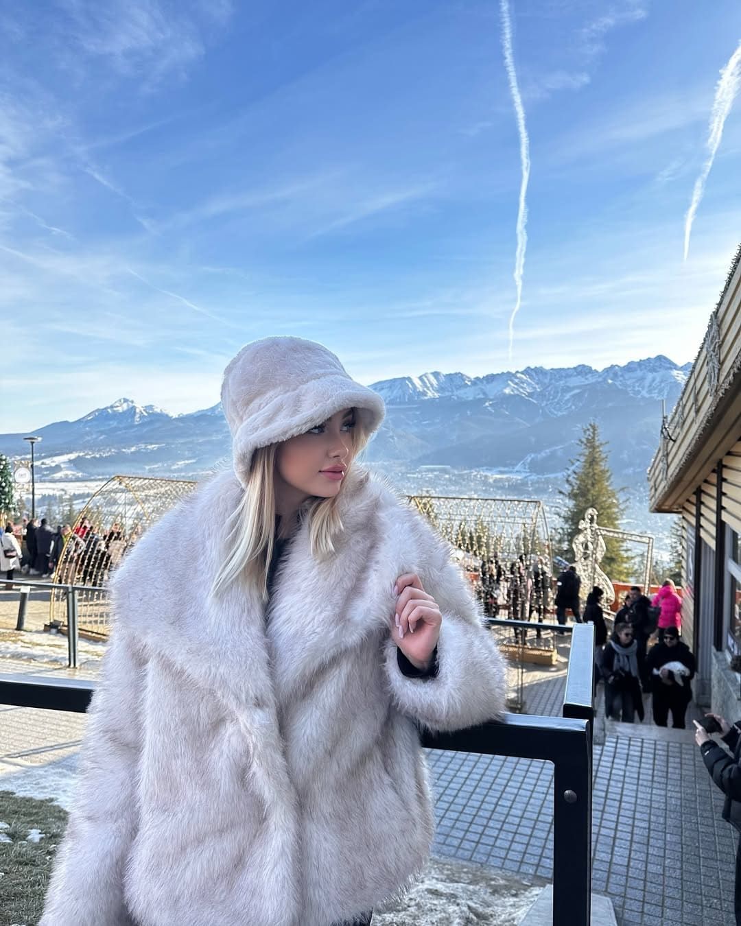 A blonde woman in a white fur coat and bucket hat rests her hand on a balcony railing overlooking a snowy town.