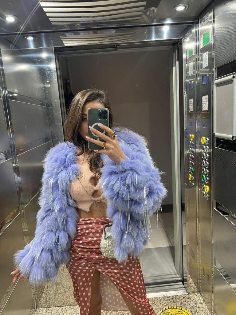Mirror selfie in an elevator showing a woman in a lavender fur coat, beige cut-out top, and red polka-dot pants holding a smartphone.