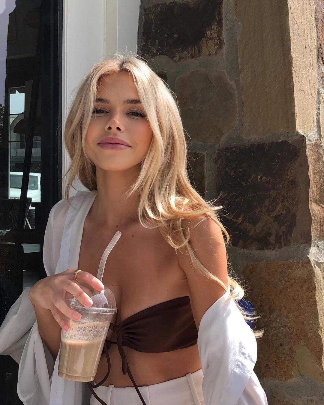 A blonde woman stands by a stone wall in a brown top and white bottoms, holding a clear cup with a straw.