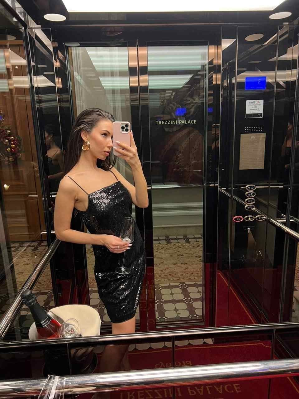 A woman in a black sequined slip dress holds a wine glass in an elevator, with a champagne bottle in an ice bucket nearby.