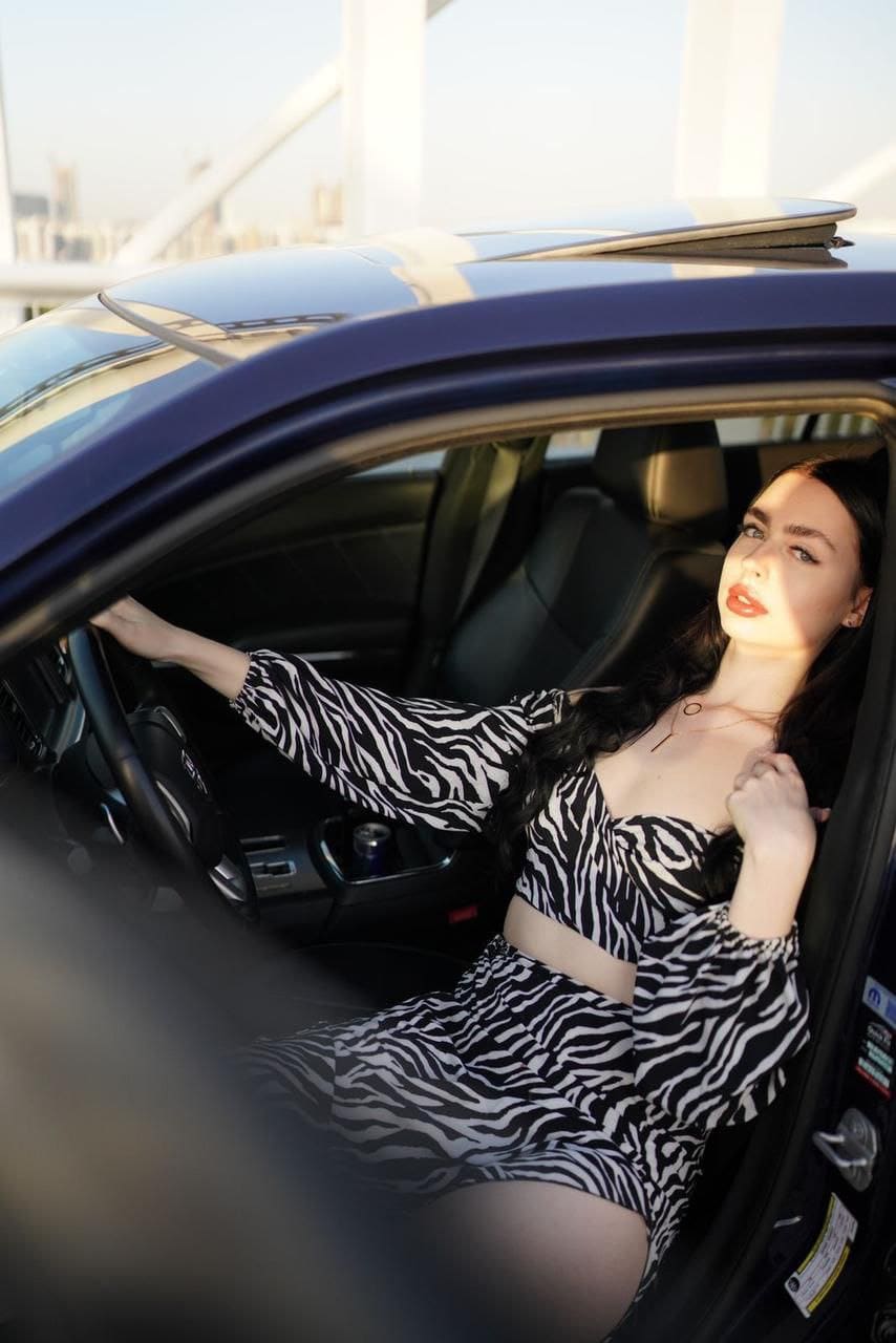 A woman in a zebra-print two-piece outfit sits in a car driver's seat, one hand on the wheel, sunlight illuminating her face.