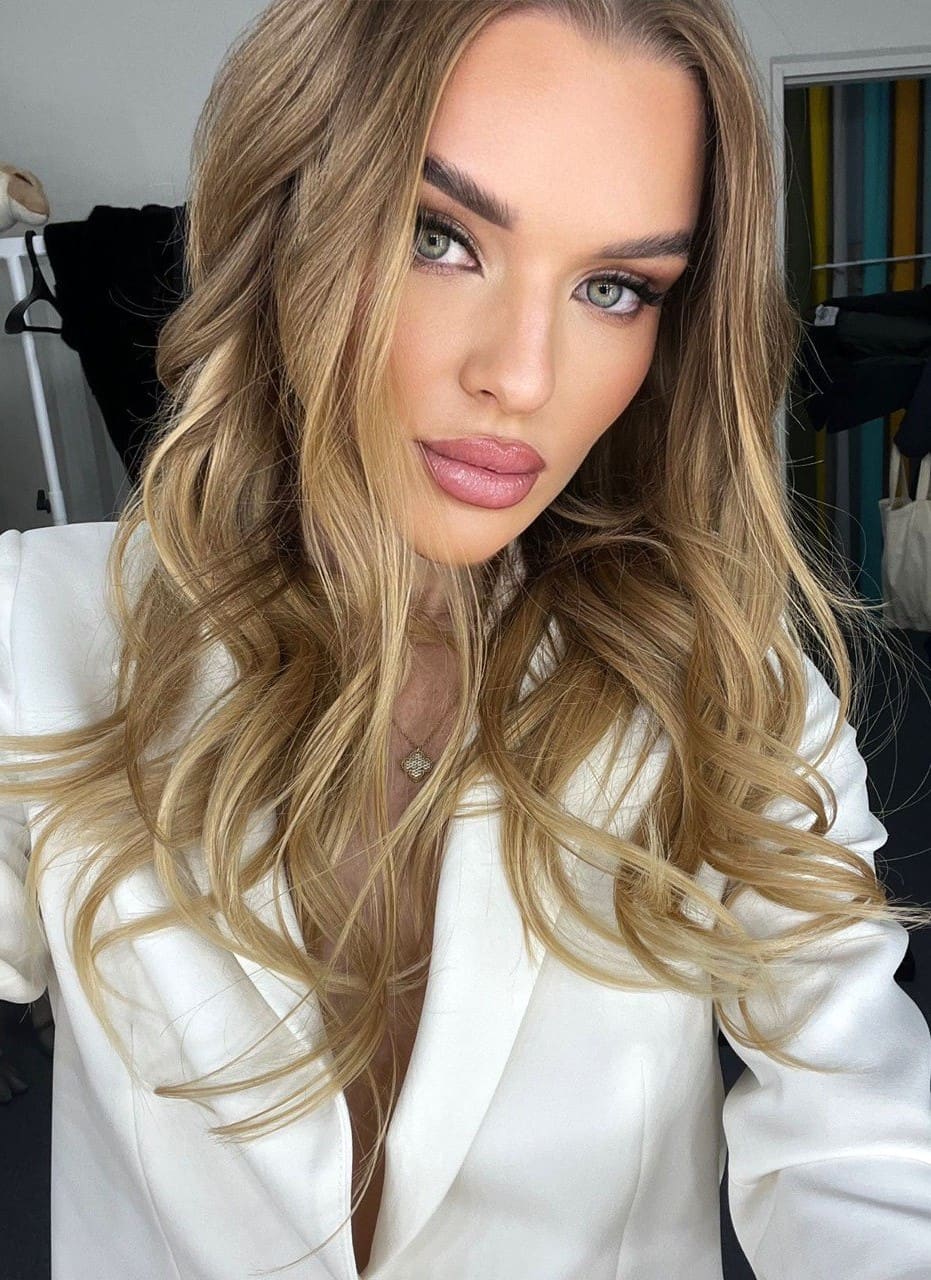 A close-up selfie of a blonde woman in a white blazer with a deep V-neck and pendant necklace, with clothing racks visible in the indoor background.