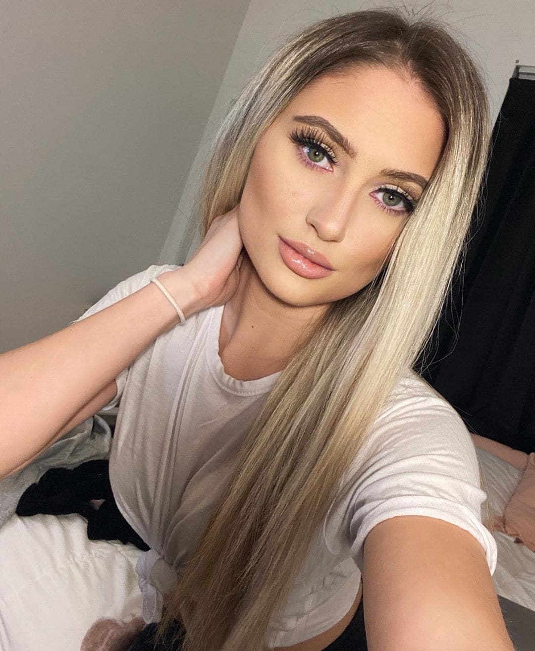 A blonde woman takes a selfie in a white t-shirt, one hand on her neck, in a bedroom with a bed and dark curtain visible behind her.