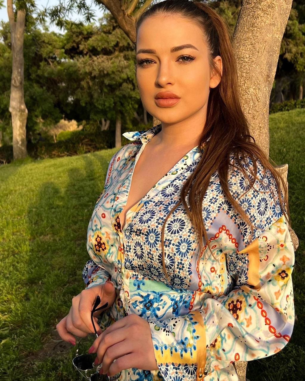 Sasha stands outdoors in a grassy area, wearing a floral blouse with a deep V-neck and holding sunglasses in her hands.