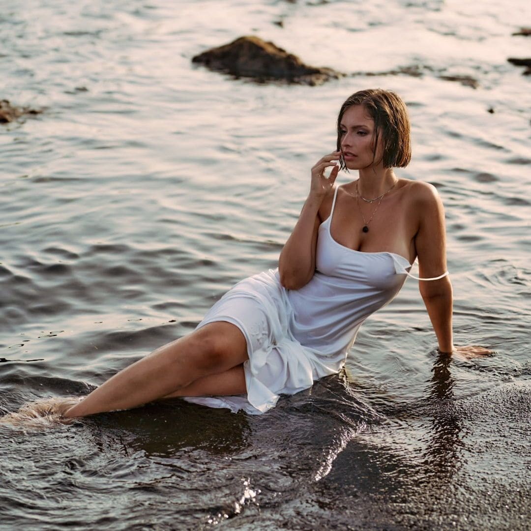 Sitting in shallow seawater, she wears a clinging white slip dress, one hand raised to her face, rocks in the background.