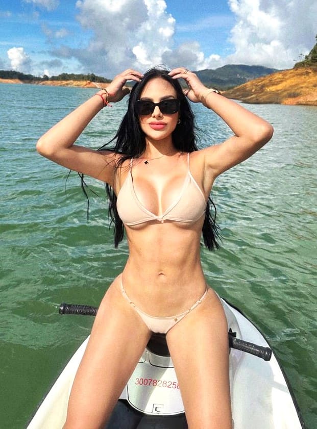Sitting on a white watercraft in a lake wearing a light bikini and sunglasses, with hands raised near her head against hills.