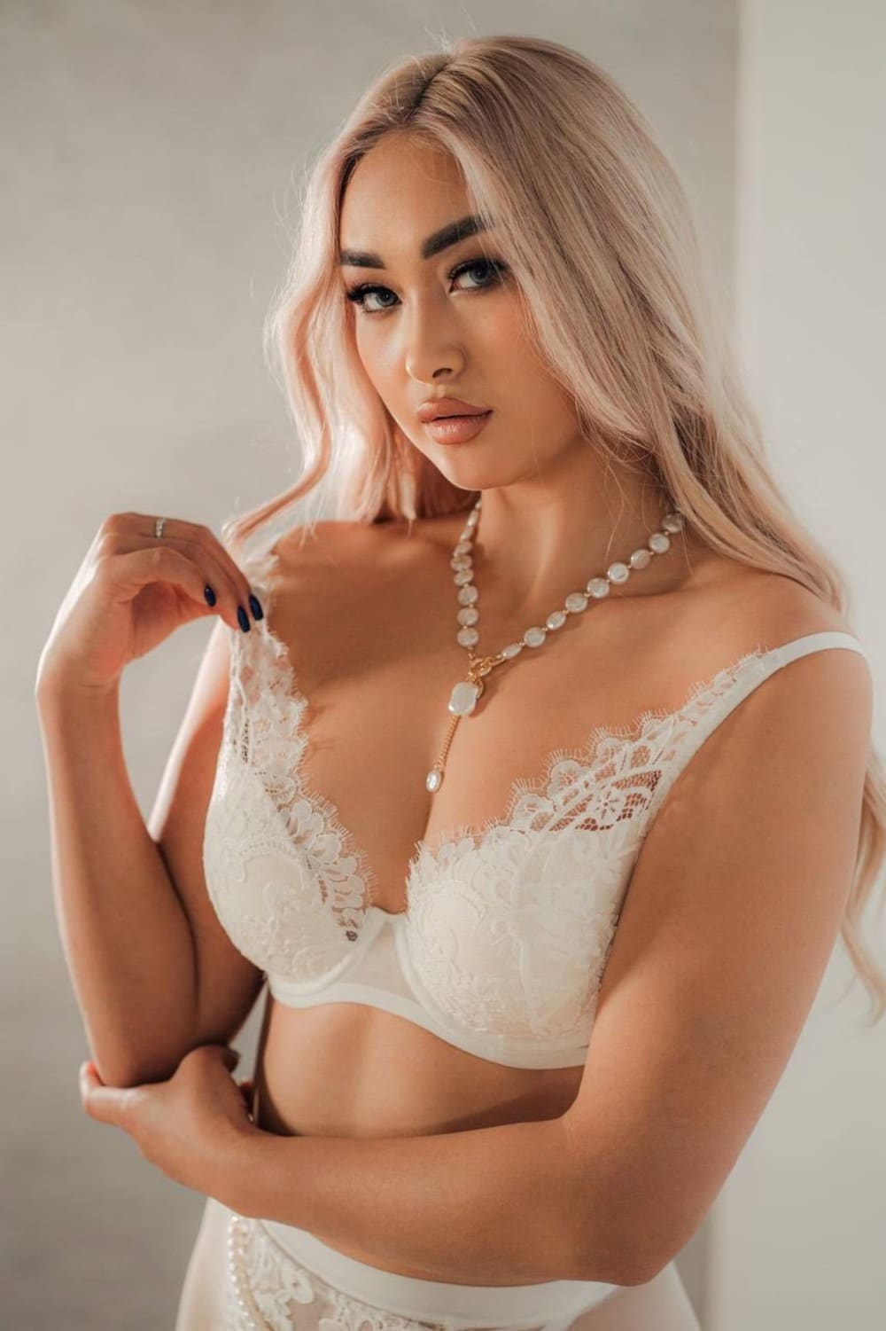 A woman stands against a plain light background in a white lace bra and matching bottoms, wearing a pearl necklace with one hand raised.